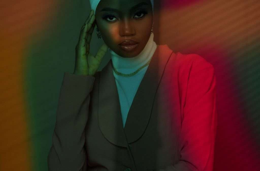 Black woman in head covering and suit bathed in multicolored light.
